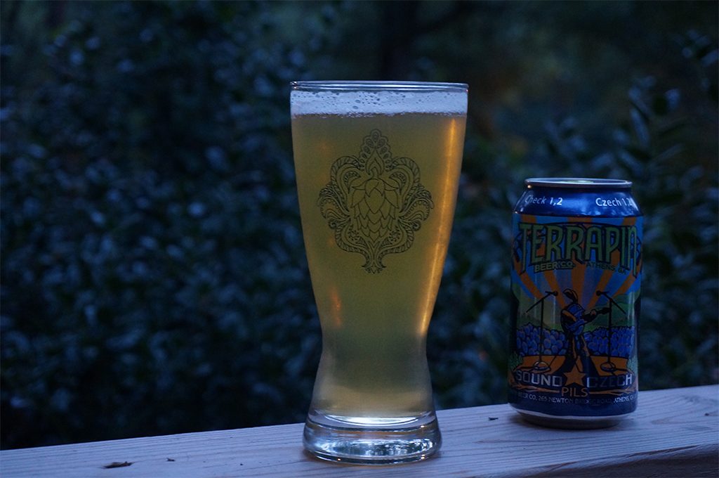 A glass of Sound Czech Pilsner neck to a can of the same on a porch railing.