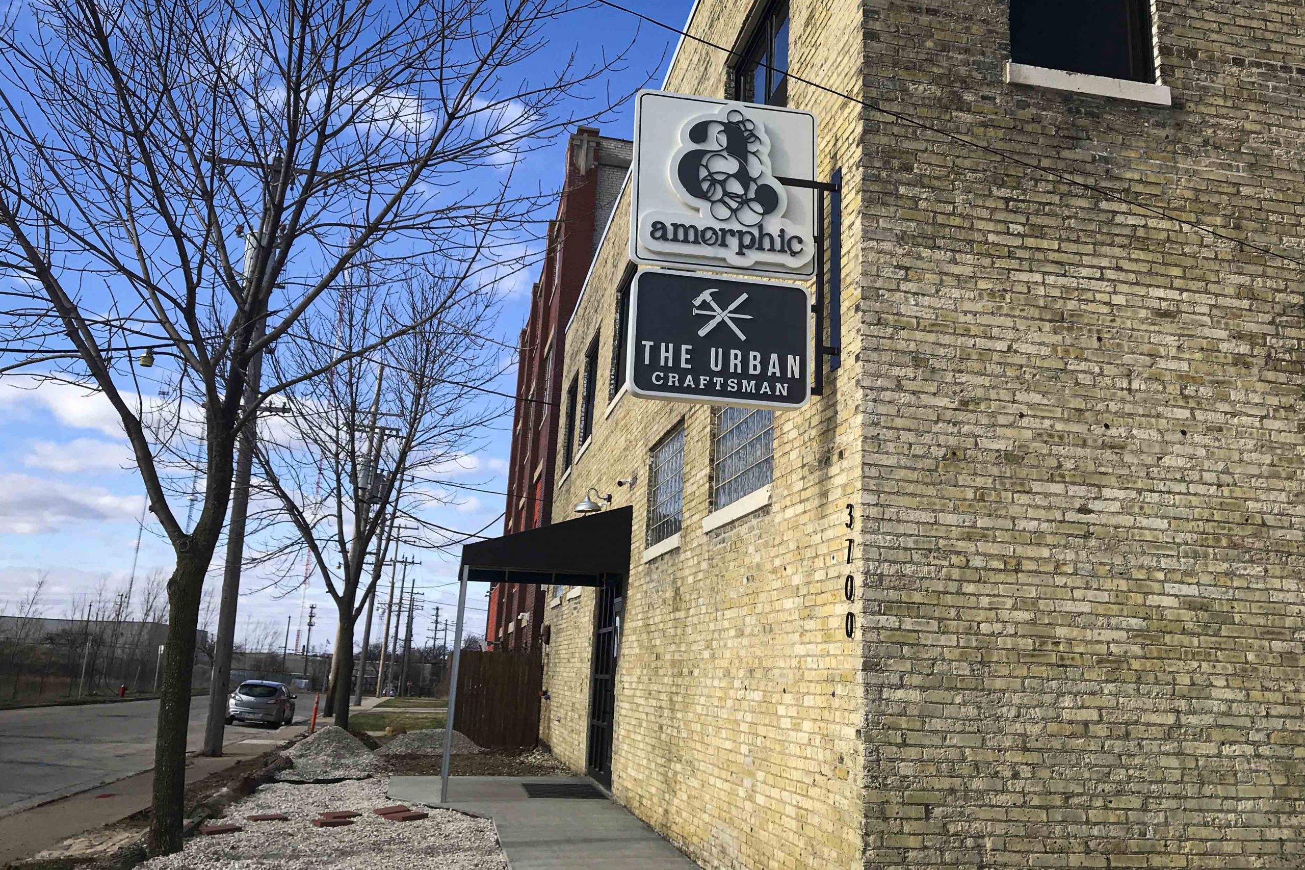 A weathered brick exterior building stands against a blue sky, with a hanging sign denoting the location of Amorphic Beer. 