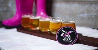 A flight of five beers sits in front of a pair of hot pink boots, and a magnet with the Pink Boots Society Collaboration Brew Day logo
