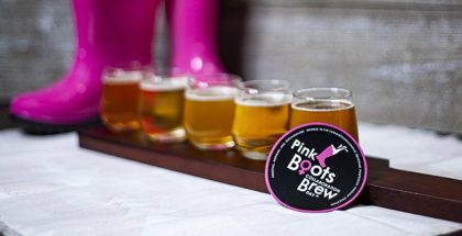 A flight of five beers sits in front of a pair of hot pink boots, and a magnet with the Pink Boots Society Collaboration Brew Day logo