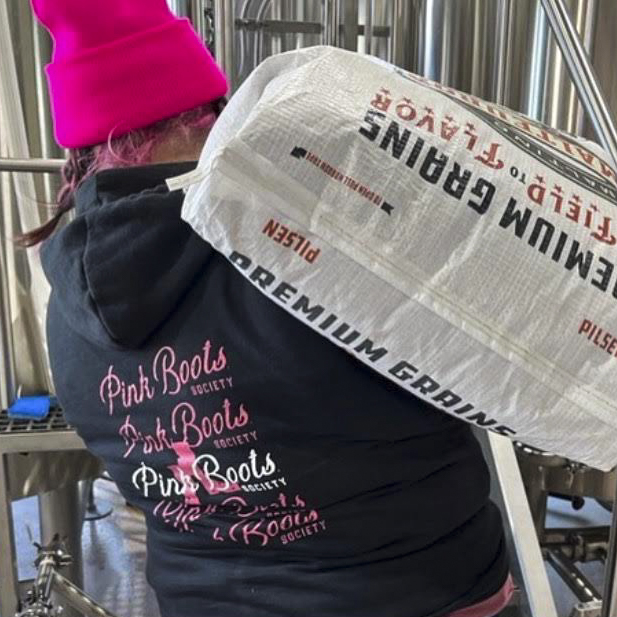 A woman wearing a Pink Boots Society sweatshirt and hot pink hat hoists a large bag of Malteurop Pilsen malt over her shoulder.