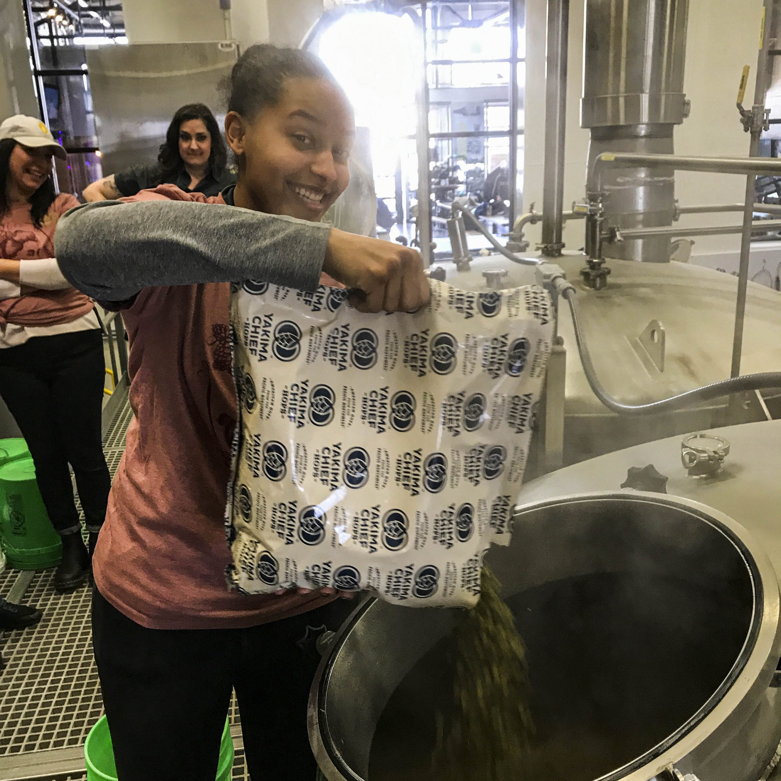 A smiling brown-skinned woman shakes a bag of hops into a whirlpool vessel while making Octavia white IPA.
