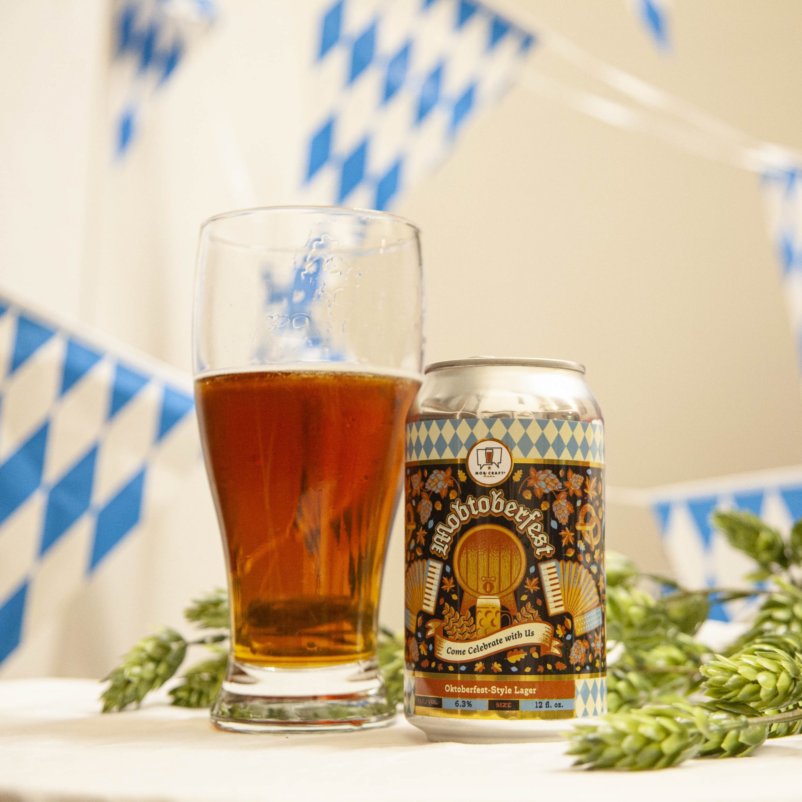 MobCraft's Mobtoberfest is poured into a pilsner glass amidst hop cones and blue and white bunting. 