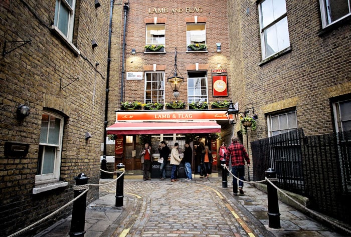 A photo of a pub, with people lingering outdoors. The street leading up to it is cobblestone, and it's between tall, potentially residential, buildings.