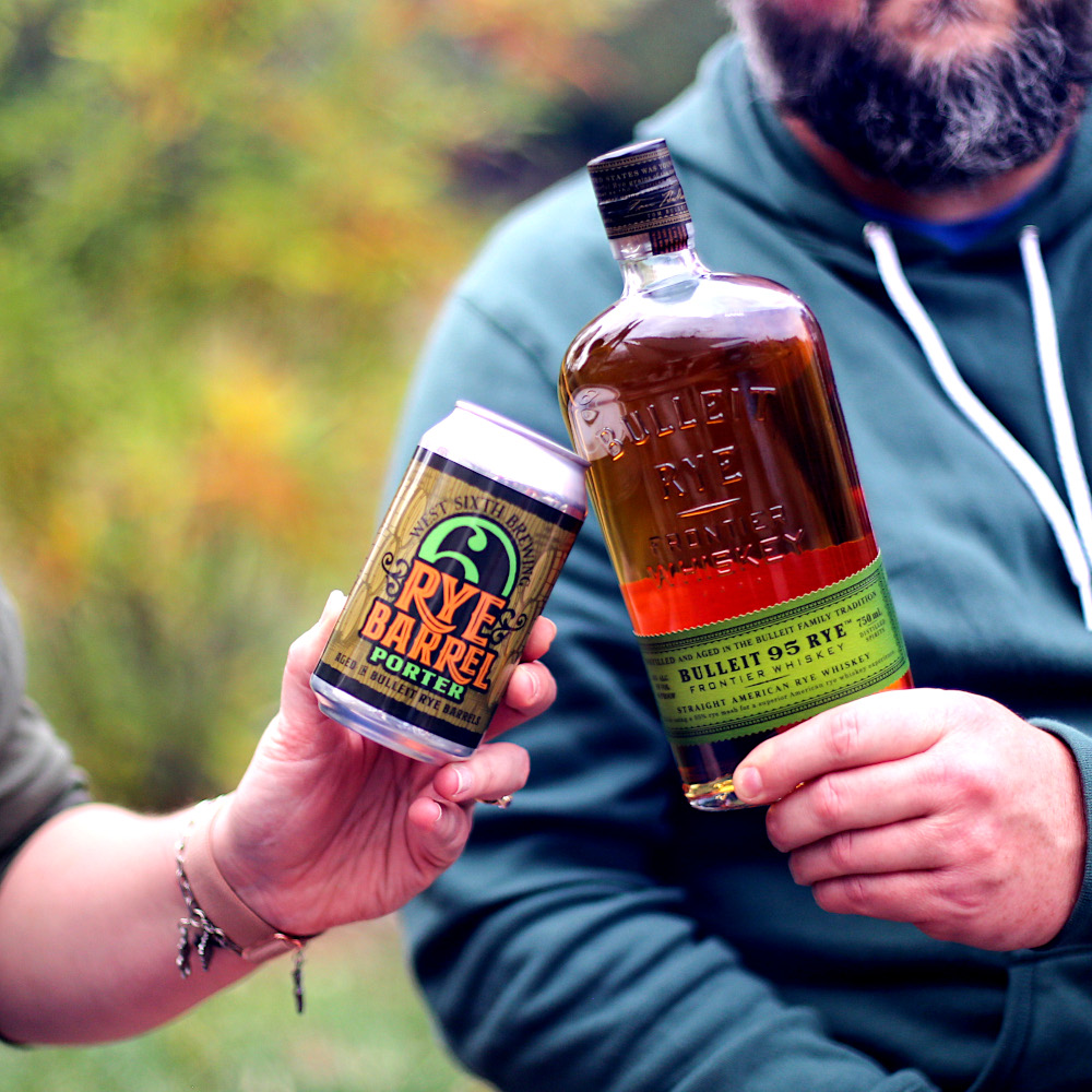 A person holding a can of West Sixth Brewing's Rye Barrel Porter touches it to a bottle of Bulleit 95 Rye held by another person.