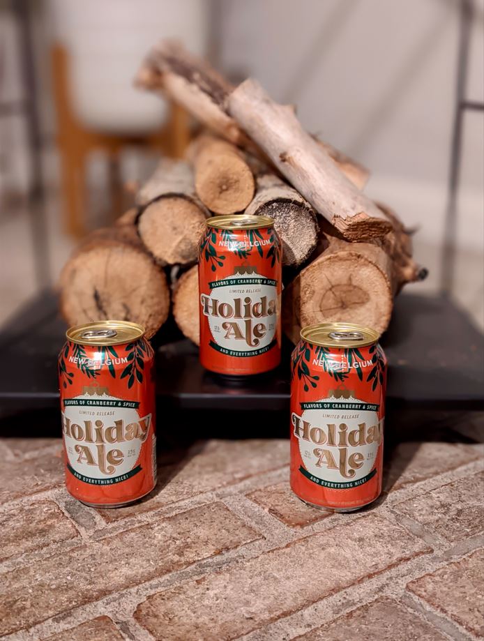 Three cans of New Belgium Holiday Ale in front of a stack of firewood on a brick hearth.