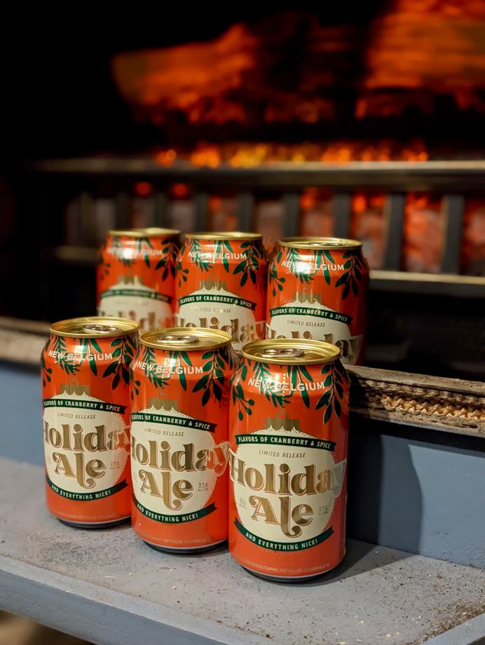 Six cans of New Belgium holiday ale in front of a cheerfully glowing fireplace grate.