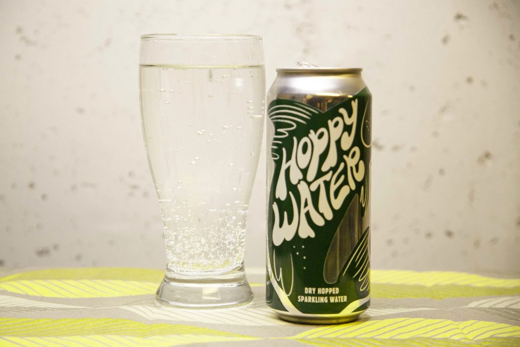 A bubbly, clear glass of Badger State Hoppy Water sits next to an empty fish-emblazoned can. 