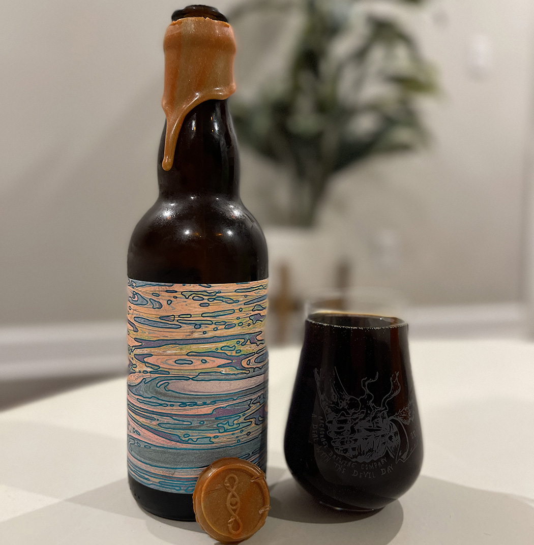 A wax-dipped bottle of beer with an abstract label alongside a full stemless glass. The beer is dark. There's a plant in the background.