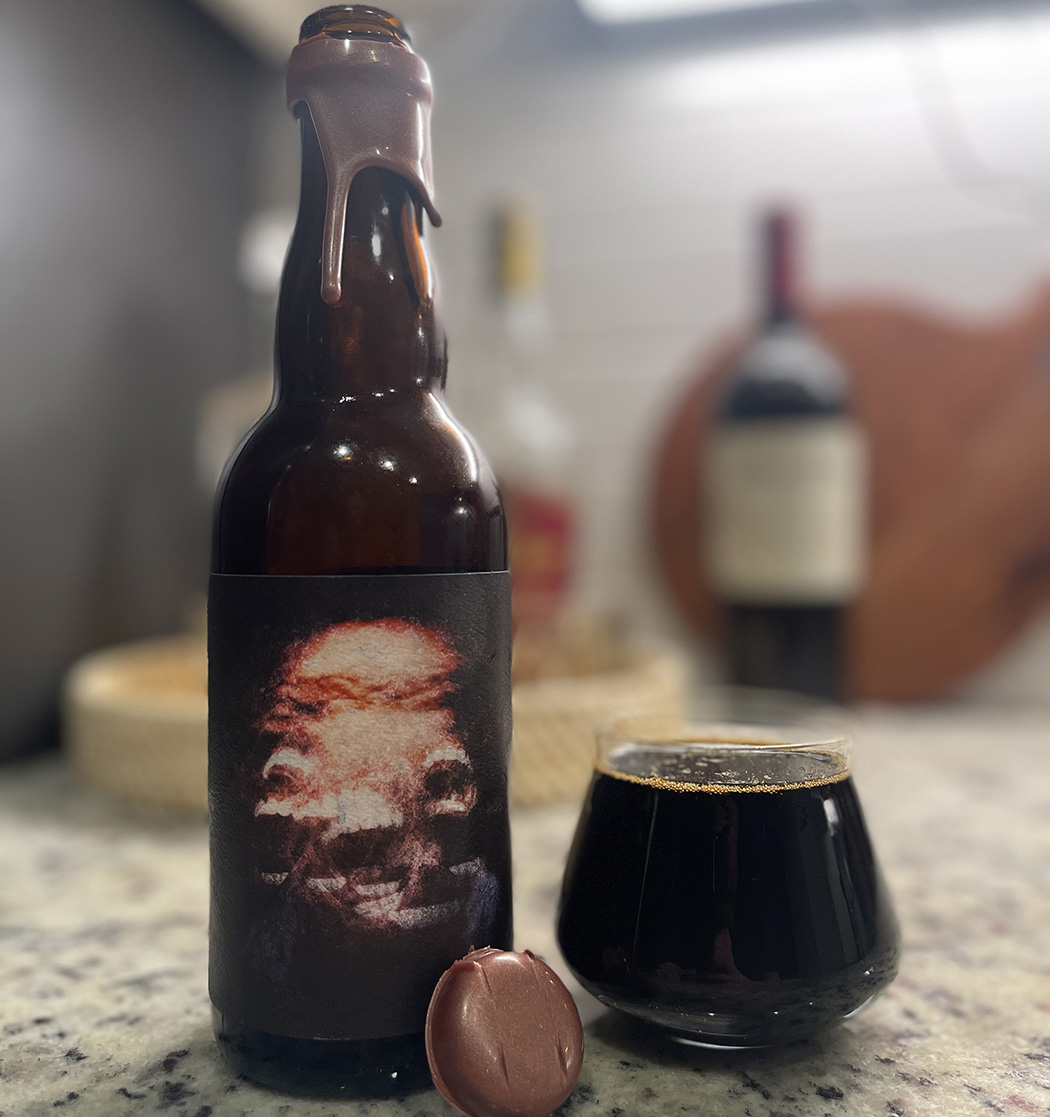 A wax-dipped bottle of beer with a label of an abstract face and teeth next to a full, dark, stemless glass of beer.