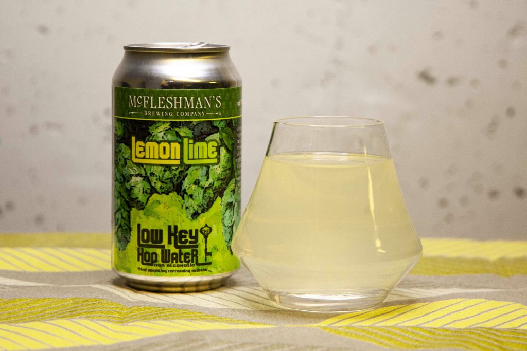 A can of McFleshman's Low Key Hop Water sits next to a glass of cloudy, yellow tinted liquid