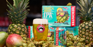 A glass of golden beer and a can in front of a six-pack box of Deschutes Brewery's Tropical Fresh IPA, surrounded by fresh pineapple and mango.