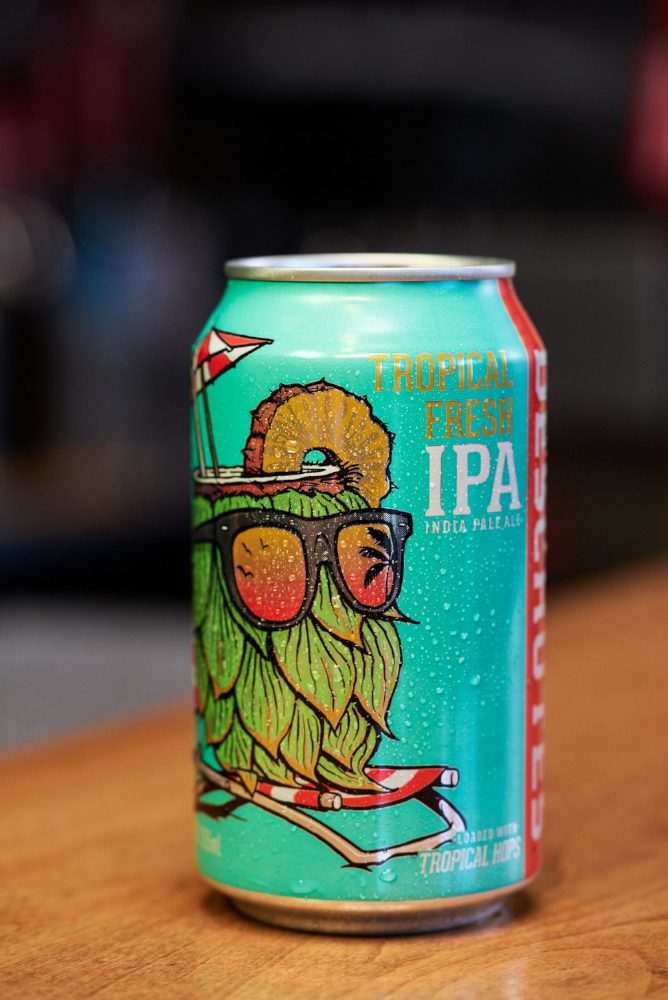 A can of Deschutes Brewery's Tropical Fresh IPA on a wooden surface. The whimsical can art features a hop on a beach chair.