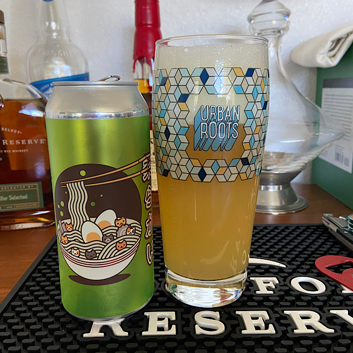 A can of Urban Roots Brewing and Smokehouse's Pile of Kittens on a bar mat next to a full glass of hazy beer with a substantial head in front of liquor bottles and a decanter.