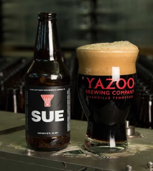 A dark beer bottle with a dark label and the simple word 'Sue' next to a glass of dark beer with a foamy head.