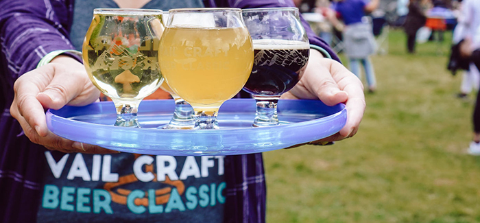 A pair of hands holds a tray of three full beer glasses outdoors.